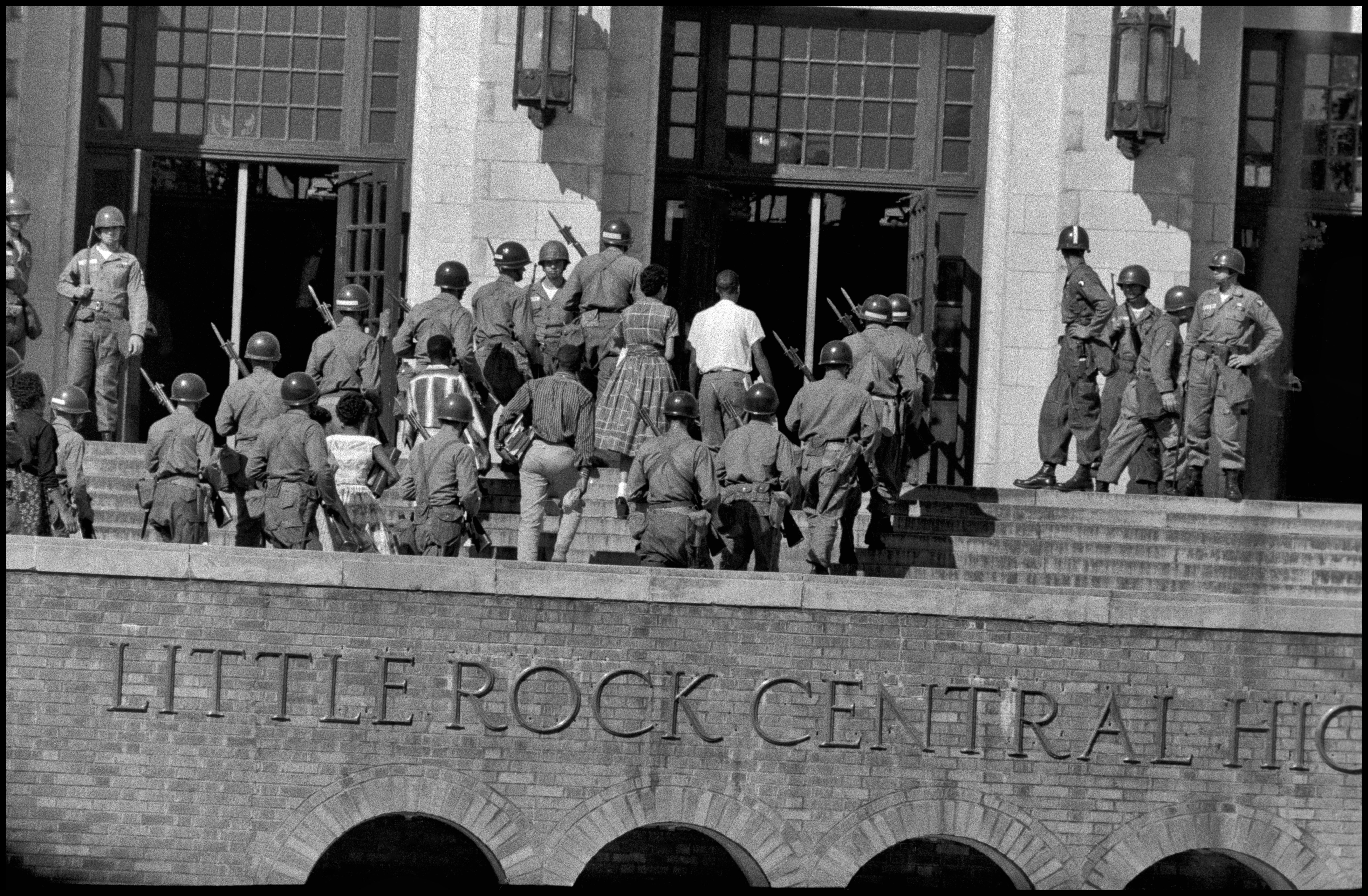 Black teenagers walking up a flight of steps into a building, surrounded by solidiers carrying guns